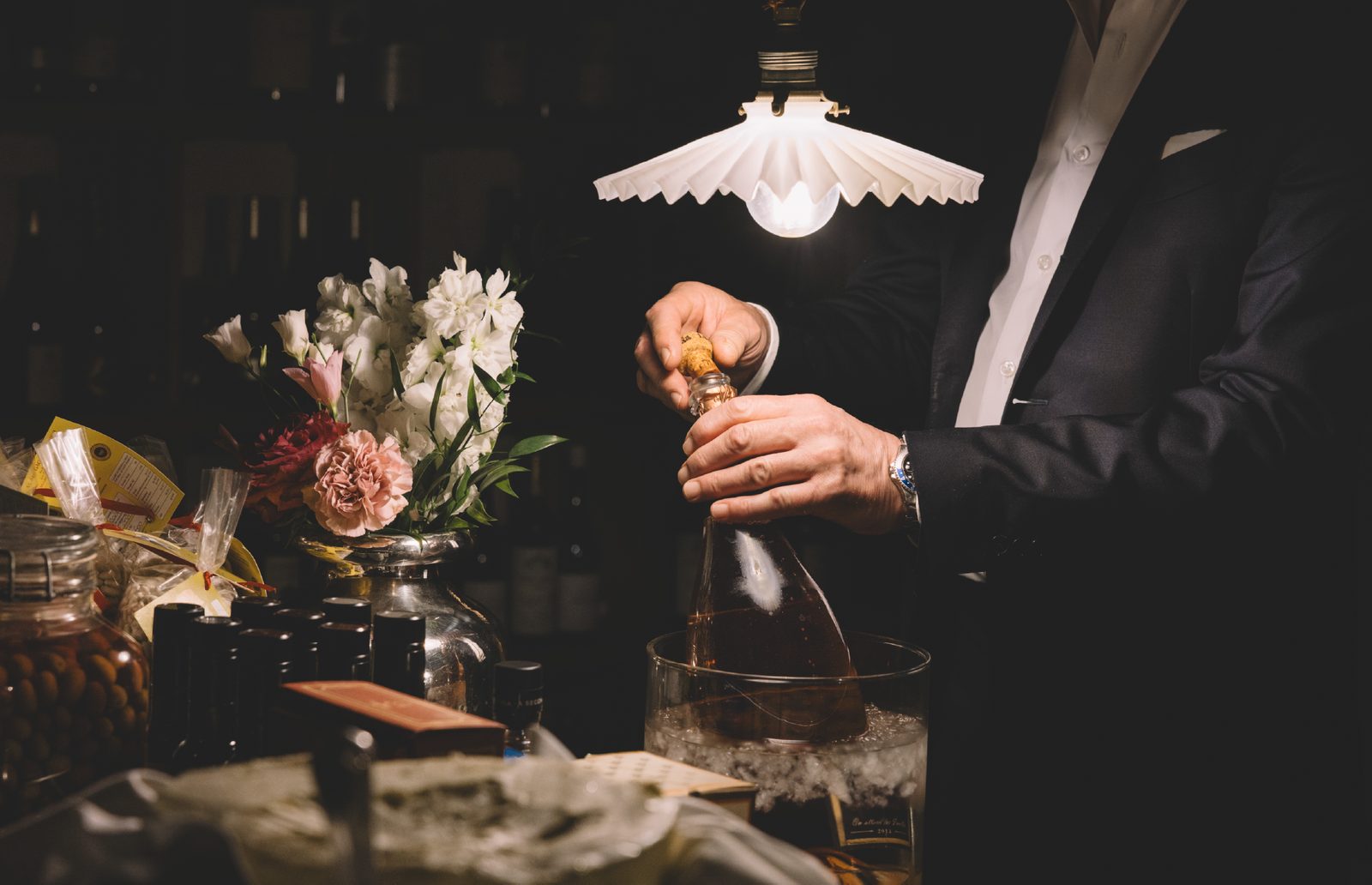 Un cameriere stappa una bottiglia in un secchiello di ghiaccio, tra fiori e prodotti, sotto una lampada nella cantina della Brughiera