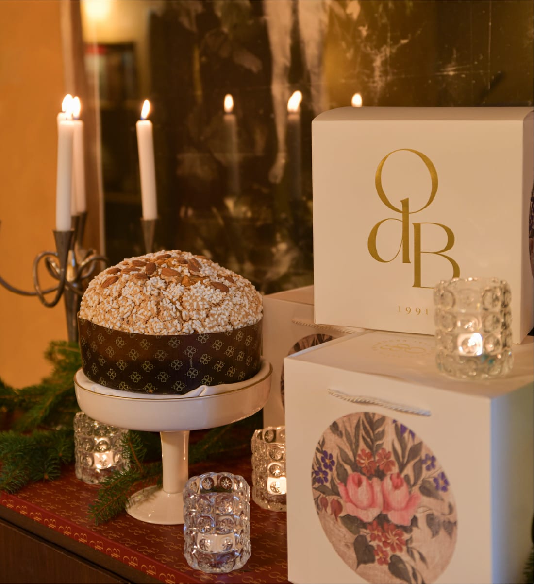 Panettone su alzata accanto a scatole con il monogramma OdB e candele accese