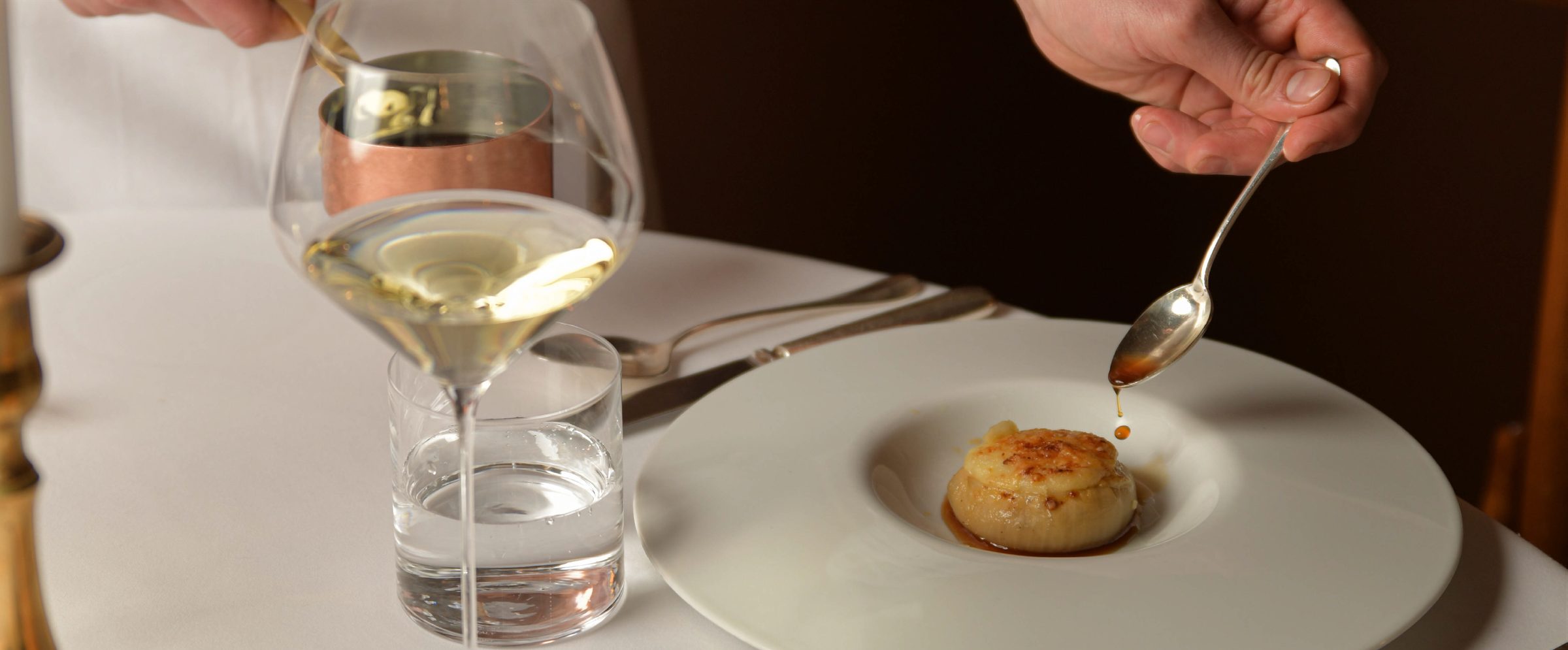 Un cucchiaio versa la salsa su una cipolla ripiena gratinata, accanto a un calice di vino bianco